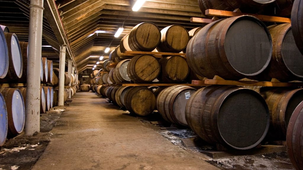 Rows of large wooden barrels stacked on racks inside a dimly lit warehouse with a concrete floor and exposed beams—the ideal setting for those curious about wholesale liquor license requirements.